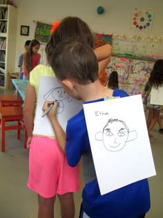 two children are drawing faces on paper in a room with other children and one child is holding a sign