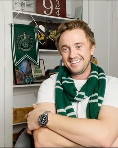 a man with his arms crossed wearing a green scarf and white t - shirt, sitting in front of a bookshelf