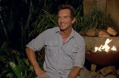 a man sitting in front of a fire pit smiling at the camera with his hands on his hips