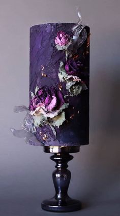 a purple cake with flowers on it is sitting on a black stand against a gray wall