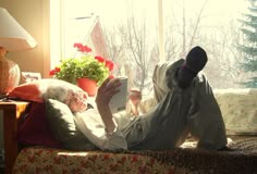 an elderly woman reading a book while laying on the couch in front of a window