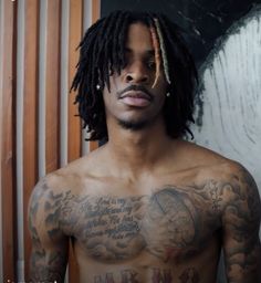 a shirtless man with dreadlocks standing in front of a wall and looking at the camera