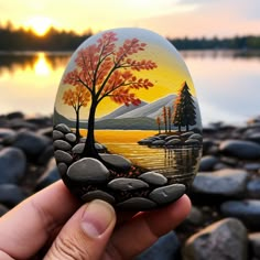 a hand holding up a painted rock with trees and rocks in front of the water