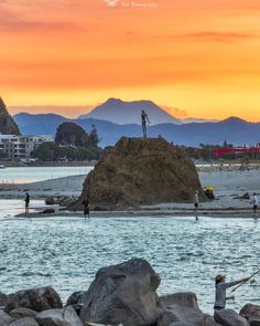 some people are fishing in the water near rocks and mountains at sunset, with one person standing on top of a rock