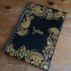 a black and gold book with jewels on the cover sitting on a wooden table next to a pen