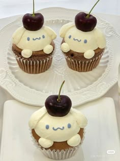 three cupcakes with white frosting and two cherries on top are sitting on a plate