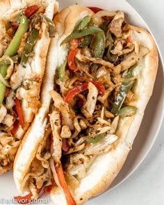 two hot dogs on buns with peppers, onions and other toppings sitting on a white plate