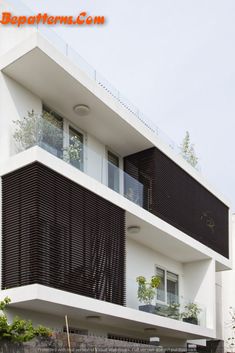 a white building with black shutters and balconies on the top floor is surrounded by greenery