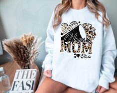 ♥LAST DAY FOR CHRISTMAS DELIVERY - 12/10/2024 Show your pride as a cheer mom with this trendy and customizable sweatshirt! Featuring bold leopard print "Cheer Mom" letters, a stylish megaphone and pom-pom design, this sweatshirt is perfect for showing off your spirit.  Add your child's name for a personalized touch that makes this cozy sweater truly unique. Whether you're cheering from the stands or attending competitions, this sweater is a must-have for moms who love supporting their cheerleaders in style! Ideal for game days, competitions, and gifting to proud cheer moms. Please note: Colors may appear less intense once printed due to the nature of the printing process. ♥ PRODUCTION TIME: 1-5 days (Usually 2-3 days) ♥ SHIPPING TIME: 2-5 days (Usually 3 days) ♥ PRODUCT DESCRIPTION: Gildan Mom Letters, Cheer Mom, Color Run, Mom Sweatshirt, Workout Sweatshirt, Cozy Sweater, Christmas Delivery, Sweater Women, Cozy Sweaters