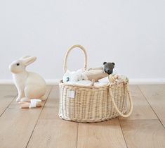 a white rabbit sitting next to a wicker basket filled with baby items on top of a wooden floor