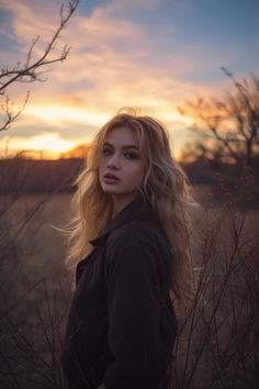 a woman standing in tall grass with the sun setting behind her and looking off into the distance