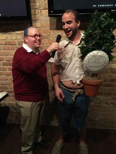 two men standing next to each other in front of a brick wall and one holding a potted plant