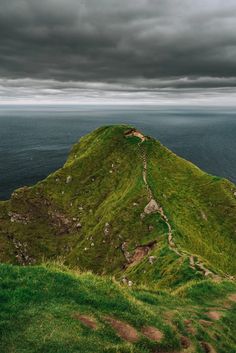 a grassy hill with a path leading to the top and ocean in the background under a cloudy sky