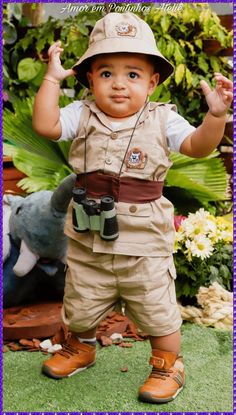 a little boy wearing a safari outfit and holding his hands up to the side while standing in front of some flowers