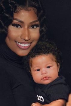 a woman holding a baby wearing a black shirt and smiling at the camera with her hands on her chest