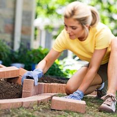 a woman in yellow shirt laying bricks on the ground with blue gloves and gardening gloves