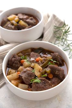 two white bowls filled with beef stew on top of a marble counter next to silverware