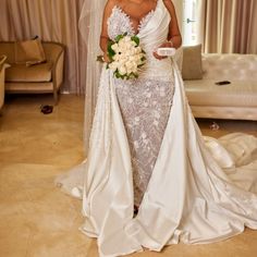 a woman in a wedding dress holding a bouquet and looking at the camera while standing next to a couch