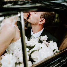 a bride and groom kissing in the back seat of a black car with white flowers