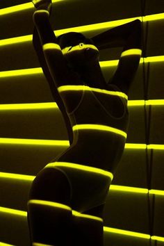 a woman standing in front of a window covered in yellow blinds with her back turned to the camera