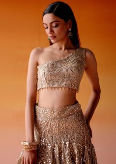 The lehenga is adorned with intricate mirrorwork and golden embellishments paired with an one-shoulder blouse in asymmetrical design, completed with mirrorwork detailing. Glamorous Gold Choli With Mirror Work, Glamorous Festive Choli With Mirror Work, Party Wear Pre-draped Saree With Gota Work For Reception, Glamorous Embellished Sets For Navratri, Glamorous Saree-style Lehenga With Mirror Work, Gold Pre-draped Saree With Mirror Work For Evening, Glamorous Choli With Mirror Work For Diwali, Glamorous Diwali Choli With Mirror Work, Pre-draped Saree With Gota Work For Reception