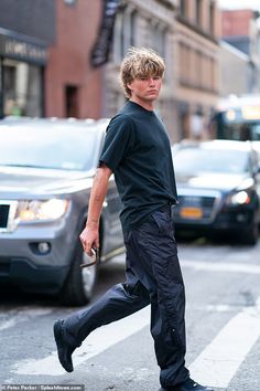 a man walking across the street in front of some parked cars and buildings, wearing all black