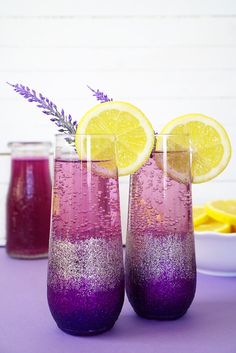 two glasses filled with purple and yellow liquid next to sliced lemons on a table