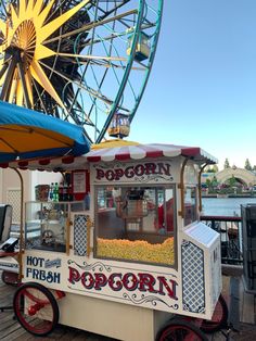 a popcorn cart sitting on top of a wooden deck next to a ferris wheel and water