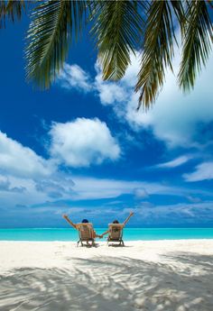 two people sitting in beach chairs under palm trees on the white sand and blue water