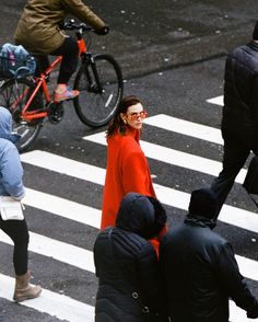 a woman in an orange coat is crossing the street with other people and bicycles behind her