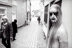 a man with long hair and makeup on walking down the street in front of some people