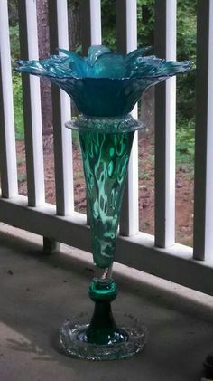 a green glass vase sitting on top of a table next to a white fence and trees