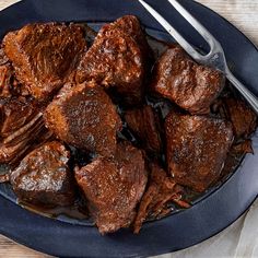 a blue plate topped with beef and potatoes next to a fork on top of a wooden table