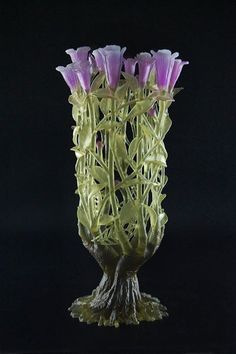 a glass vase with purple flowers in it on a black tableclothed surface, against a dark background