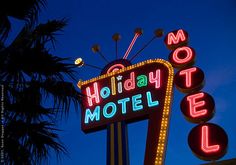 a motel sign lit up at night with palm trees in the foreground and blue sky behind it