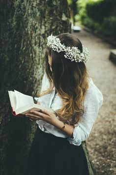 a woman is standing by a tree and reading a book with a flower crown on her head