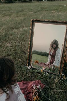 a woman sitting on the ground in front of a mirror with flowers and grass around her