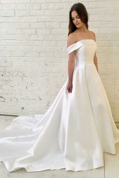 a woman standing in front of a brick wall wearing a white wedding dress with an off the shoulder neckline