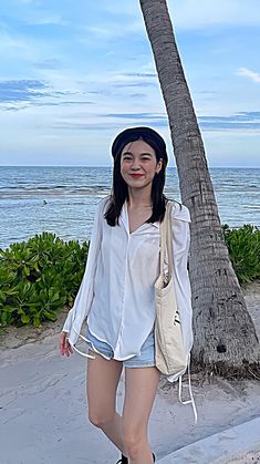 a woman standing next to a palm tree on top of a sandy beach with the ocean in the background