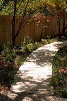 a wooden path in the middle of a garden with trees and shrubs around it, leading to a fence