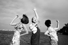 three women standing next to each other in front of the ocean with their hands up