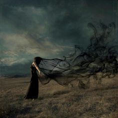 a woman standing in a field with her long black dress flowing over her head and hair blowing in the wind