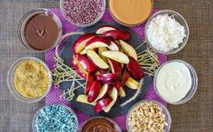 an assortment of fruits and nuts in bowls on a table with chocolate, peanut butter, apple slices, almonds, sprinkles, and other toppings