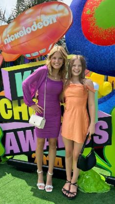 two women standing next to each other in front of a colorful sign with balloons on it