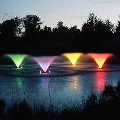 four fountains are lit up in the night sky over water with trees and bushes behind them