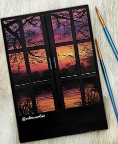 the painting is next to two paintbrushes and an open window with sunset outside