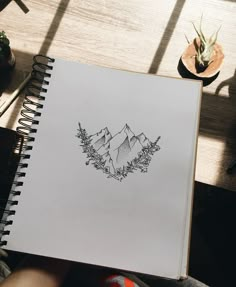 someone holding up a notebook with a drawing of mountains and pine trees on the cover