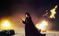 a woman dressed in black holding a knife and standing next to fire on the ground