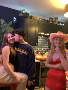 three people standing in a kitchen posing for the camera with one woman wearing a pink hat