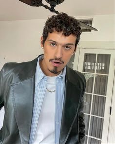 a man with curly hair wearing a black leather jacket and white t - shirt standing in front of a ceiling fan